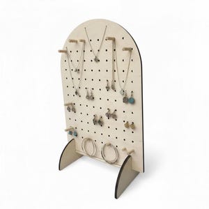 Puede incluir: Un expositor de joyas de madera clara con forma arqueada y dise&ntilde;o de tablero perforado con numerosos agujeros peque&ntilde;os. El soporte incluye varias clavijas de madera horizontales para colgar collares y pendientes que se muestran en &eacute;l.