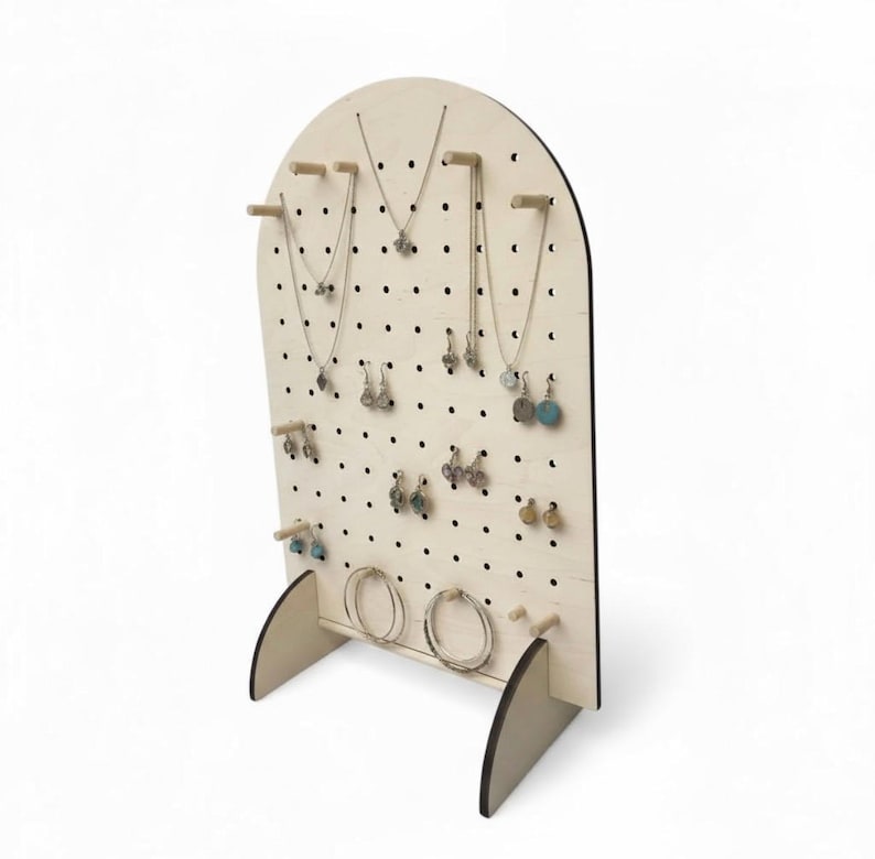 Puede incluir: Un expositor de joyas de madera clara con forma arqueada y dise&ntilde;o de tablero perforado con numerosos agujeros peque&ntilde;os. El soporte incluye varias clavijas de madera horizontales para colgar collares y pendientes que se muestran en &eacute;l.