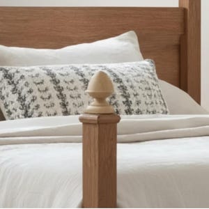 May include: A wooden bed frame with a light-colored headboard and footboard. A decorative pillow with a black and white pattern rests on the bed. The bed is covered with white bedding. A wooden finial is in the foreground.