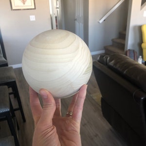 May include: A close-up of a light wood sphere, with a smooth, slightly textured surface. The sphere is being held in a hand, with a gold ring on the index finger.