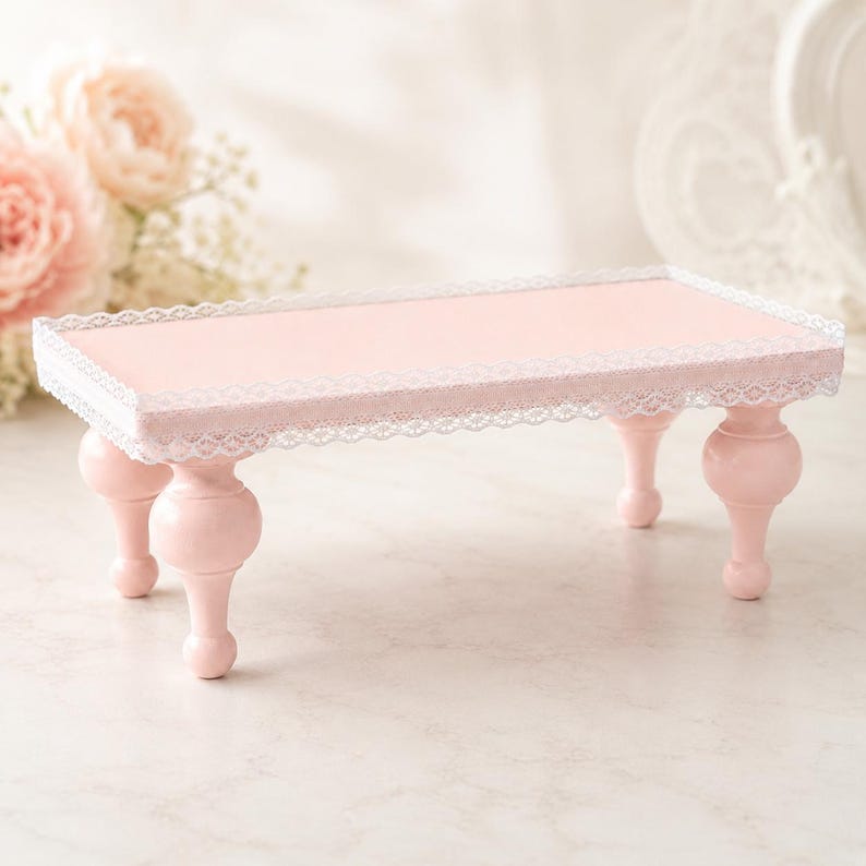 Op de afbeelding: Een kleine, rechthoekige, roze serveerschaal met een delicate witte kanten rand. De schaal wordt ondersteund door vier afgeronde, roze poten. De schaal wordt waarschijnlijk gebruikt voor het presenteren van kleine items of het serveren van lekkernijen.