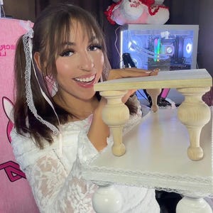 Op de afbeelding: Een kleine, witte decoratieve tafel met gedraaide houten poten en een kanten rand. De tafel heeft een licht houten blad en wordt ondersteund door gedraaide houten poten. De tafel is ontworpen voor decoratieve doeleinden.
