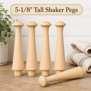 May include: Five light-colored wooden Shaker pegs, each measuring 5-1/8 inches tall. Four pegs stand vertically in a row, while one peg lies horizontally in the foreground. The pegs feature a smooth, tapered design with a rounded top and a base.