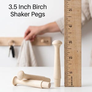 May include: Light-colored birch wood Shaker pegs with a rounded top and a tapered base. The pegs measure 3.5 inches in length as indicated by the wooden ruler placed vertically next to them. The text 3.5 Inch Birch Shaker Pegs is visible.