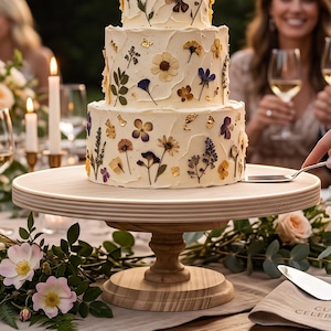 May include: A three-tiered white frosted cake decorated with pressed flowers and gold leaf flakes, resting on a round wooden pedestal stand. The cake features various botanical elements including small purple, yellow, and white blossoms.