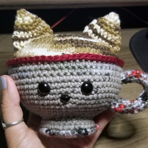 May include: A handmade crochet cat mug in gray, brown, and red. The mug features a cat face design with black eyes and a handle. The cat ears are brown and beige. The mug is held in a hand.