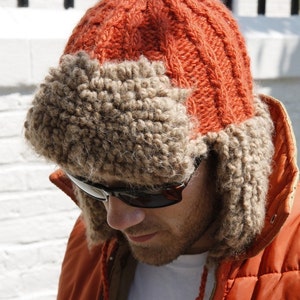 May include: A man wearing a brown and orange knitted hat with ear flaps and a matching orange puffer jacket.