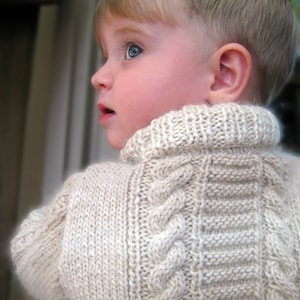 Pode incluir: Uma criança pequena vestindo um suéter de tricô cor creme com um padrão de tricô trançado. O suéter tem gola alta e mangas compridas.