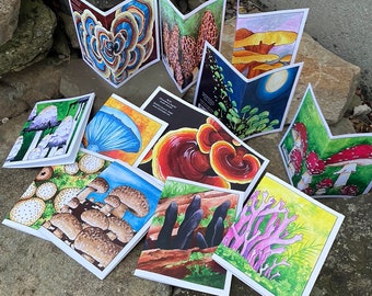 May include: A collection of folded greeting cards featuring colorful mushroom illustrations. The cards display various fungi, including puffballs, morels, and bracket fungi, with detailed artwork and vibrant hues. The cards are arranged on a stone surface.