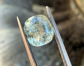 May include: A light green, cushion-cut gemstone held by silver tweezers. The gem has internal fractures and a multifaceted surface, reflecting light. The background is blurred, focusing attention on the gem's details. The tweezers are positioned to display the gem.