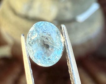 May include: A light blue, oval-shaped gemstone held by silver tweezers. The gem has internal inclusions and a translucent quality, with a subtle, watery appearance. The background is blurred, suggesting a close-up shot of a precious stone.