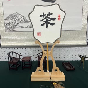 May include: A white folding fan with black calligraphy that reads "茶" (tea) on it. The fan is on a wooden stand with a brown tassel.