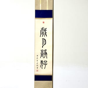 Könnte beinhalten: Eine traditionelle japanische Schriftrolle mit schwarzer Kalligraphie auf beigefarbenem Hintergrund. Die Schriftrolle hat einen blauen und goldenen Rand und ist auf einem Holzstab montiert. Die Kalligraphie lautet "前 功" und bedeutet "früheres Verdienst".