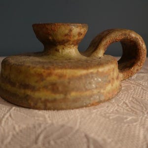 Può includere: Un portacandele in ceramica fatto a mano con manico. Il portacandele è un mix di tonalità marroni, gialle e verdi, con una superficie strutturata. Il manico ha una forma arrotondata e il portacandele poggia su una tovaglia di pizzo bianco.