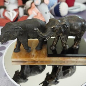 Op de afbeelding: Twee bronzen olifantenbeeldjes, één donker en één lichter, staan naast elkaar op een spiegelend oppervlak. De olifanten hebben omhoog geheven slurf en gedetailleerde kenmerken. Ze staan op een marmeren en zwarte basis, die eronder weerspiegelt.