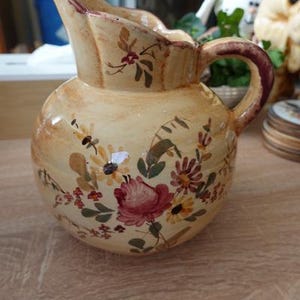 Op de afbeelding: Een keramische kan met een crèmekleurig glazuur en een handgeschilderd bloemmotief met roze rozen, gele madeliefjes en groene bladeren. De kan heeft een ronde vorm, een geschulpte rand en een gebogen handvat met een donkerbruine rand.