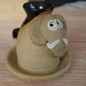 Peut inclure: Un brûleur d'huile en céramique en forme d'éléphant fantaisiste. L'éléphant est beige avec des défenses blanches et de grands yeux de dessin animé. Le brûleur a un dessus noir et une petite base en forme de plat. Un objet décoratif pour l'aromathérapie.