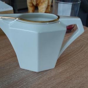 Può includere: Una lattiera bianca di forma geometrica con bordo e manico dorati. La lattiera ha un beccuccio e un manico. Il design è moderno ed elegante, adatta per servire caffè o tè. Misura circa 15 cm di lunghezza.