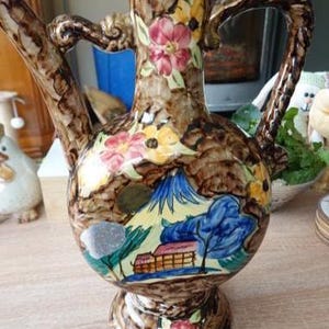 Può includere: Una brocca in ceramica con smalto marrone maculato, caratterizzata da motivi floreali dipinti a mano e una scena paesaggistica con una montagna, alberi e un edificio. Il recipiente ha un corpo largo e rotondo, collo stretto, manico e beccuccio.