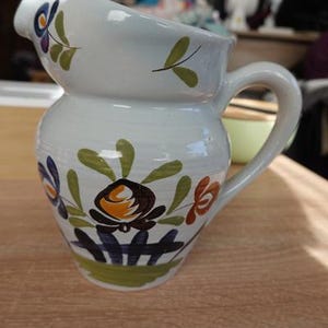 Pode incluir: Jarro de cerâmica branca com bico curvo e corpo arredondado. O jarro é decorado com desenhos florais coloridos em laranja, azul e verde. Possui uma alça curva e está sobre uma superfície de madeira.