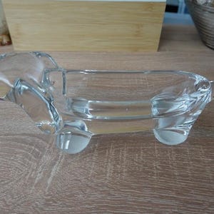 Op de afbeelding: Een heldere, teckelvormige schaal van glas. De glazen hond heeft een lang lichaam, korte poten en een gebogen staart. De schaal is ontworpen om kleine items in te bewaren en staat op een houten oppervlak.
