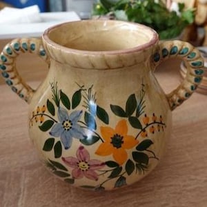 Op de afbeelding: Een crèmekleurige keramische vaas met twee handvatten met een touwstructuur. Het lichaam is voorzien van handgeschilderde bloemmotieven in blauw, oranje en roze, vergezeld van groene bladeren en kleine gele bessen. De rand heeft een touwachtig reliëf.