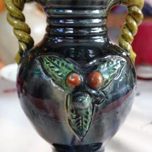 Könnte beinhalten: Eine dunkelblaue Keramikvase mit zwei gedrehten Seilhenkeln. Die Vase hat ein grünes und orangefarbenes Blumendesign mit einer erhabenen Textur.