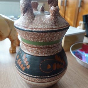 May include: A ceramic water jug with a twisted handle. The jug features a speckled brown and tan body with a black band at the base, decorated with orange leaf designs. A green stripe encircles the jug.