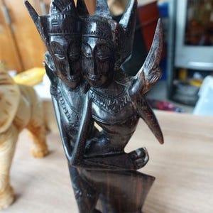 Op de afbeelding: Een donkerbruin houten beeldhouwwerk dat twee figuren afbeeldt met sierlijke hoofdtooien en gestileerde kenmerken. Het beeldhouwwerk heeft een gladde, gepolijste afwerking en staat op een houten oppervlak. De figuren zijn verweven, wat wijst op een nauwe relatie.