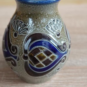 Op de afbeelding: Een kleine keramische vaas met een gespikkelde, bruinachtige achtergrond en een blauwe rand. De vaas heeft een decoratief patroon in tinten blauw, bruin en bruin. Het ontwerp bevat bloem- en geometrische elementen.