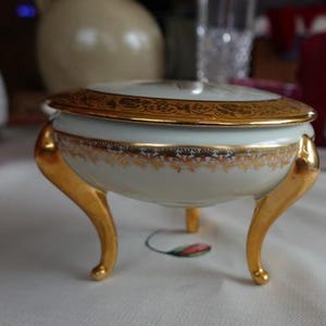 Könnte beinhalten: Eine weiße Porzellan-Schmuckdose mit goldenem Deckel und Verzierungen. Die Dose steht auf drei goldenen Beinen.