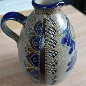 May include: A ceramic pitcher with a blue handle and rim, featuring a decorative pattern of blue and brown floral designs. The pitcher has a spout for pouring and a textured, speckled surface.