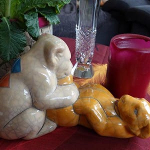 Könnte beinhalten: Zwei Keramik-Tierfiguren, eine weiße mit bunten geometrischen Mustern und eine gelbe, sind auf einer roten Oberfläche zusammengestellt. Im Hintergrund befinden sich eine rote Kerze und eine klare Glasvase.