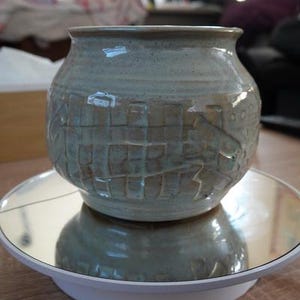 May include: A light blue ceramic vase with a textured surface and carved design. The round vase has a wide opening and sits on a white and mirrored surface. The vase is approximately 18 cm in diameter.