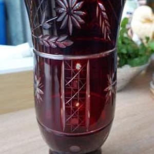 Può includere: Un vaso in vetro rosso intenso con dettagli in vetro molato. La parte superiore presenta motivi floreali e di grano incisi, mentre il corpo inferiore mostra pannelli verticali con un motivo a griglia e accenti a forma di stella.