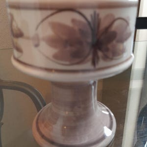 Pode incluir: Um pedestal de cerâmica branca com um design floral castanho. O pedestal tem um topo redondo e uma base larga.
