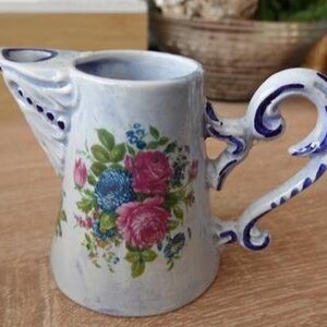 Può includere: Una lattiera in ceramica con finitura smaltata azzurra, decorata con un bouquet di rose rosa e fiori blu sul davanti. La lattiera presenta un manico ornamentale lavorato e un beccuccio dalla forma particolare con dettagli dipinti in blu.