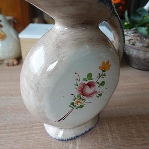 Può includere: Brocca in ceramica color crema con corpo bulboso e beccuccio curvo. Presenta un motivo floreale con una rosa rosa e fiori gialli. Il bordo e la base sono rifiniti in blu. Un oggetto decorativo per la cucina.