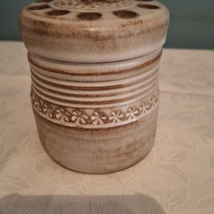 May include: A white ceramic jar with a brown lid. The lid has a decorative design with small holes. The jar has a brown and white striped design with a row of small flowers.