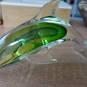 Op de afbeelding: Een dolfijnfiguur van helder glas met een levendige groene kern ingesloten in het transparante lichaam. Het beeldje is afgebeeld in een springende houding met een gebogen rugvin, een spitse snuit en een gladde afwerking over het geheel.