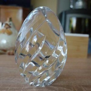 May include: A clear, egg-shaped glass ornament with a carved, spiral design. The glass reflects light, creating a shimmering effect. The object is on a wooden surface.