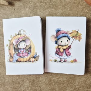 May include: Two white notebooks featuring illustrations of mice. The left notebook shows a mouse sitting inside a large pumpkin. The right notebook depicts a mouse wearing a blue hat and yellow scarf, holding a colorful autumn leaf umbrella.
