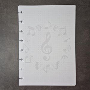 May include: A white notebook with a musical theme. The notebook features a light grey design of musical notes and a treble clef. The pages have a dotted grid pattern. The notebook is bound with black rings.