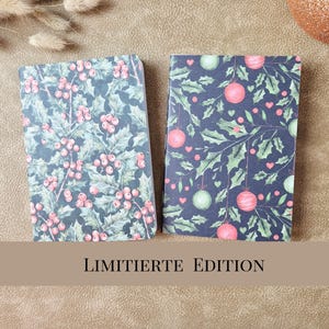 May include: Two rectangular notebooks with dark backgrounds. The left notebook features a pattern of red holly berries and green leaves. The right notebook displays a pattern of hanging red and green ornaments, holly leaves, and small hearts.