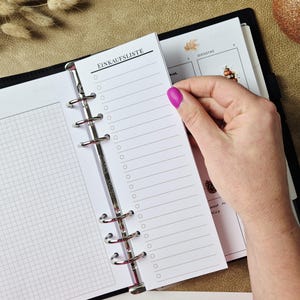 May include: A white paper insert for a ring binder featuring the text Einkaufsliste at the top and a vertical column of empty circular checkboxes with horizontal lines for writing, held within a black binder with grid paper pages.