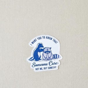 Op de afbeelding: Een witte sticker met een blauwe illustratie van een wasbeer en een pinguïn. De tekst op de sticker luidt: "I want you to know that Someone Cares, Not Me, But Someone."