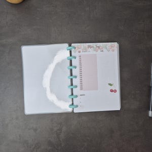 May include: Open planner with a light blue binding. The right page has a pink section for time slots and the words "Mein Tag" at the top. Cherry illustrations are at the top and bottom of the page. A pen is visible on the right.