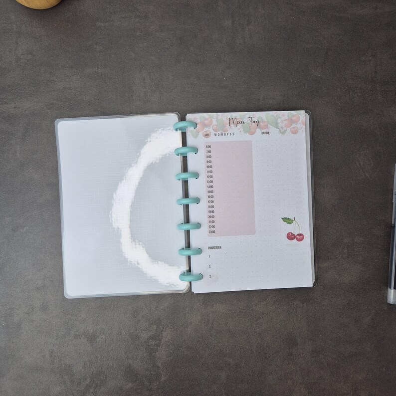 May include: Open planner with a light blue binding. The right page has a pink section for time slots and the words "Mein Tag" at the top. Cherry illustrations are at the top and bottom of the page. A pen is visible on the right.