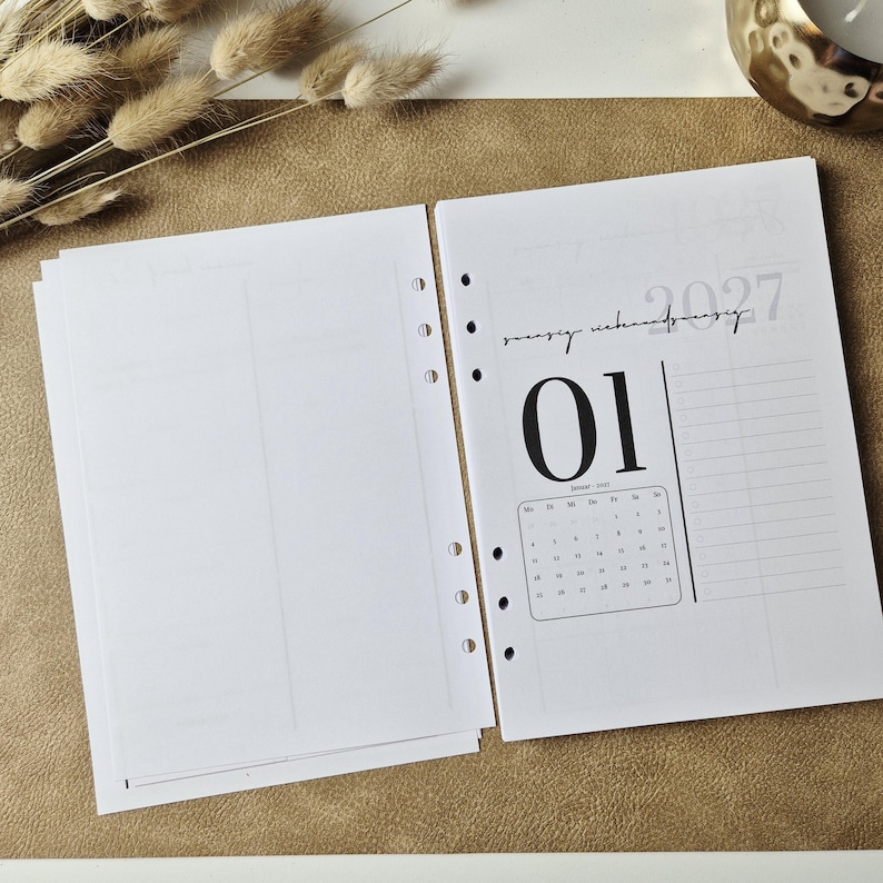 May include: A set of white planner refill pages with punched holes along the side. The right page features a 2027 calendar for January, the number 01 in large black font, and a vertical checklist. The left page is blank with a faint grid layout.
