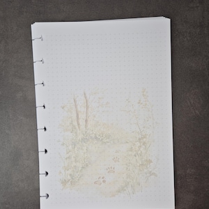 May include: A white dot grid notebook with a nature-themed illustration. The illustration features a path with paw prints, trees, and foliage in soft, muted tones. The notebook has a spiral binding on the left side.